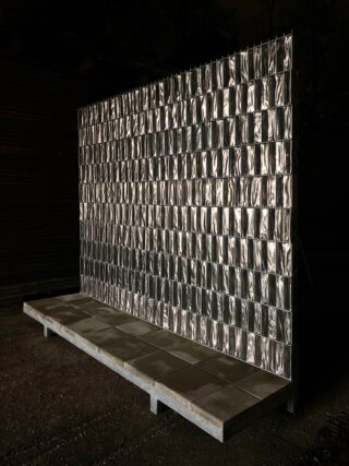 Eine metallische Gitterwand mit rechteckigen reflektierenden Platten steht nachts auf einer Betonplattform in einem schwach beleuchteten Außenbereich.