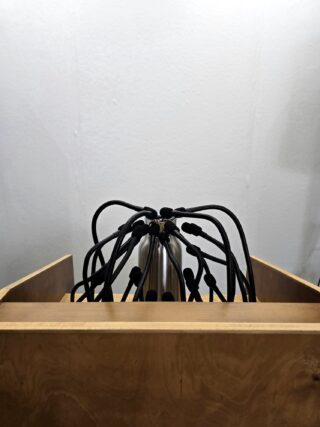 Ein silberner Zylinder mit mehreren daran befestigten schwarzen Kabeln steht in einem Holzgehäuse vor einer schlichten weißen Wand.