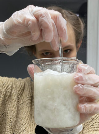 Eine Person mit Handschuhen rührt mit einem Metallstab eine trübe weiße Mischung in einem Becherglas.