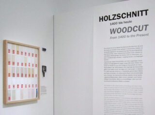 Wanddisplay mit dem Titel „HOLZSCHNITT 1400 bis heute / WOODCUT From 1400 to the Present“ mit einem gerahmten Kunstwerk und einem erklärenden Text in Deutsch und Englisch.