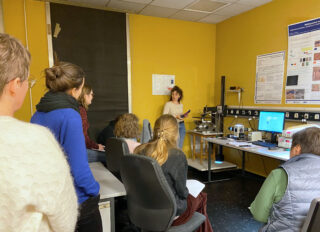 Eine Frau steht vor einem Labor mit gelben Wänden und präsentiert einer Gruppe, die in der Nähe von Computern und wissenschaftlichen Geräten sitzt und steht, Informationen.