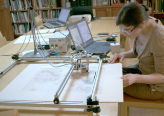 Eine Person beobachtet einen großen Scanner oder ein Plottergerät, das eine Zeichnung auf einem Tisch analysiert, im Hintergrund stehen Laptops und Bücher.