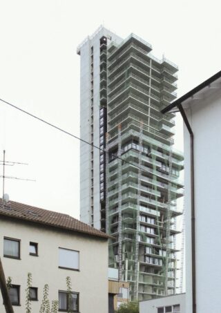 Hohes Hochhaus im Bau mit Gerüst, hinter niedrigeren Wohngebäuden an einem bewölkten Tag.