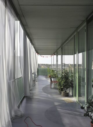 Ein langer Balkon mit grünem Geländer und Glastüren, durchsichtigen weißen Vorhängen, Topfpflanzen, einer Bank, einem Fahrrad und roten Wimpelfahnen darüber.