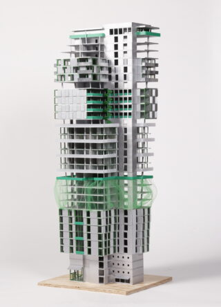 Ein hohes Architekturmodell mit grauen und grünen versetzten kubischen Formen und freiliegenden Strukturelementen, präsentiert auf einem Holzsockel vor einem schlichten Hintergrund.