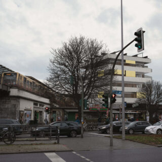 Straßenkreuzung mit an einer roten Ampel stehenden Autos, links vorbeifahrender Hochbahn, blattlosen Bäumen, modernem Gebäude im Hintergrund und bewölktem Himmel darüber.