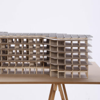 Ein maßstabsgetreues Architekturmodell eines mehrstöckigen Gebäudes mit freiliegenden Böden und Strukturelementen, ausgestellt auf einem Holztisch vor einem schlichten weißen Hintergrund.