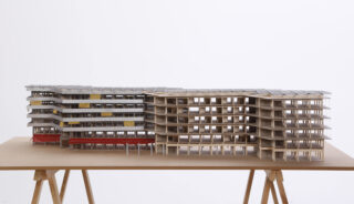 Architekturmodell eines mehrstöckigen Gebäudes mit versetzten Stockwerken, großen Fenstern und unregelmäßig gewelltem Design, ausgestellt auf einem Tisch mit Holzstützen vor einem schlichten Hintergrund.