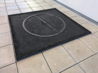 Ein quadratisches Kunstwerk auf einem gefliesten Boden zeigt einen in dunkle Erde gezeichneten Kreis, über dessen Durchmesser ein gerader Metallstab verläuft.