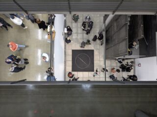 Draufsicht auf Menschen, die sich in einem Lobbybereich versammelt haben, mit einer großen schwarzen Matte, auf der ein weißer Kreis und eine diagonale Linie abgebildet sind.