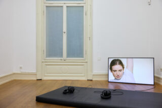 Eine flache schwarze Matte mit zwei Kopfhörern liegt auf einem Holzboden neben einem Bildschirm, auf dem eine Frau in einem weißen Oberteil zu sehen ist. Das Ganze steht vor einer weißen Wand mit einem hohen Fenster.