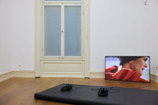 Auf dem Boden steht ein kleiner Fernseher, der die Nahaufnahme einer Person in einem roten Oberteil zeigt; zwei Kopfhörer liegen auf einer schwarzen Matte vor einem Fenster in einem spärlich möblierten Raum mit Holzboden.