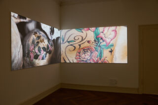 Eine digitale Projektion an zwei benachbarten Wänden zeigt Nahaufnahmen von Blumentattoos und verzierten Oberflächen in einem schwach beleuchteten Raum mit Holzboden.