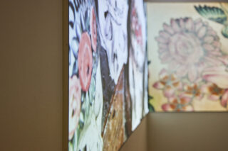 Zwei beleuchtete Kunstwerke mit floralen und abstrakten Mustern werden an benachbarten Wänden in einer Galerieumgebung ausgestellt.