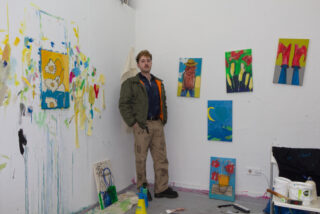 Eine Person steht in einem Kunstatelier, umgeben von farbenfrohen Gemälden an den Wänden und auf dem Boden verstreuten Utensilien.