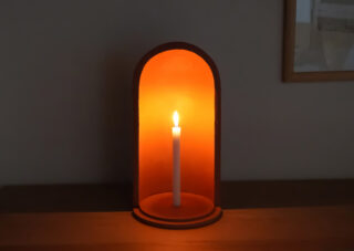 Eine einzelne brennende weiße Kerze steht aufrecht in einem gewölbten orangeroten Halter auf einer Holzoberfläche vor einer schlichten Wand.