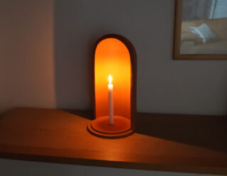 Eine brennende weiße Kerze steht aufrecht in einem gewölbten Holzhalter auf einem Holzregal und wirft ein sanftes, warmes Licht auf die nahe Wand.