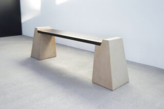 Eine minimalistische Bank mit einer dunklen Holzsitzfläche und zwei großen, trapezförmigen Betonstützen auf einem hellen Boden.