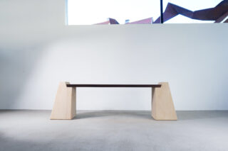 Minimalistische Bank mit dunkler Holzsitzfläche und zwei hellen, dreieckigen Betonstützen, im Innenbereich vor einer weißen Wand mit hohem Fenster aufgestellt.