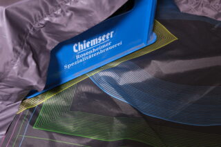 Eine blaue Kiste mit der Aufschrift „Chiemseer Rosenheimer Spezialitätenbrauerei“, teilweise bedeckt mit einem grauen und bunten Stoff mit geometrischem Muster.