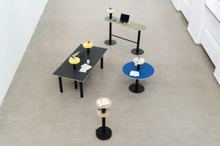 Ein minimalistischer Arbeitsbereich mit fünf Tischen in unterschiedlichen Formen und Farben, auf denen auf einem hellen Boden Gegenstände wie Notizbücher, ein Laptop, Tassen und kleine Pflanzen stehen.