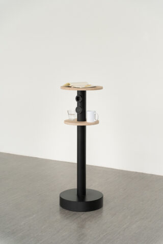 Ein kleiner, moderner Säulentisch mit zwei runden Holzregalen; auf dem oberen Regal liegen ein geschlossenes Buch und eine Brille, auf dem unteren Regal ein Glas und eine weiße Tasse.