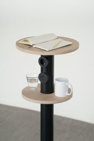 Ein kleiner, runder, zweistufiger Beistelltisch mit einem Glas Wasser, einer weißen Tasse, einem offenen Notizbuch und einem Stift. Der Tisch hat eine zentrale schwarze Stange mit integrierten Steckdosen.
