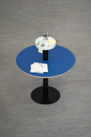 Auf einem runden Tisch mit blauer Tischplatte liegen ein offenes Notizbuch mit einem Stift und ein Tablett mit Stiften, Haftnotizen, Büroklammern und anderen Schreibwaren.