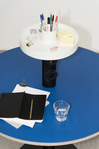 Auf einem blauen, runden Tisch liegen ein schwarzes Notizbuch mit Stift, Papier mit einer Heftklammer, ein Glas Wasser und ein Organizer mit Stiften, Haftnotizen und Büromaterial.