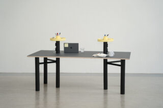 Ein minimalistischer Schreibtisch mit einem geschlossenen Laptop, einem Notizbuch, einer Tasse, einem Glas Wasser und zwei gelben Stifthaltern vor einer schlichten weißen Wand.