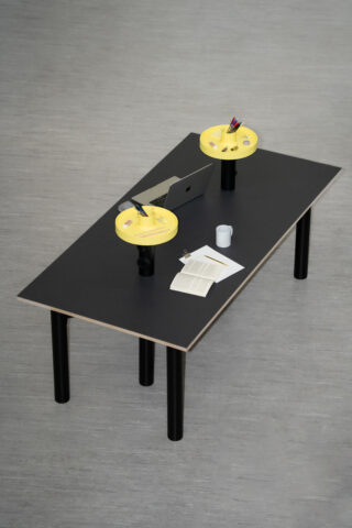 Ein schwarzer Tisch mit zwei gelben Schreibtisch-Organizern, einem Laptop, einem offenen Buch, Papieren, einem Stift und einer weißen Tasse auf einem hellgrauen Boden.