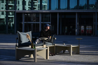 Person, die auf Gartenmöbeln vor einem modernen Glasgebäude sitzt, wobei das Sonnenlicht Schatten wirft.