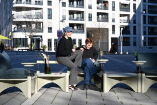 Zwei Personen sitzen im Freien auf modernen Stühlen, lachen und unterhalten sich. Einer hält ein Getränk in der Hand. Sie befinden sich in einer städtischen Umgebung mit einem weißen Gebäude im Hintergrund.