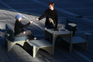 Zwei Personen stoßen an, während sie an einem modularen Gartenmöbelset auf einer gepflasterten Fläche sitzen.