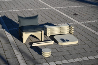 Zerlegte Gartenmöbel mit Kissen, Paneelen und Verbindungsstücken auf einer gepflasterten Fläche ausgelegt.