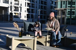 Eine Frau und ein Kind sitzen auf Gartenmöbeln. Das Kind liest ein Buch, während die Frau ein Getränk hält. Flaschen und Gläser stehen auf Tischen in der Nähe. Im Hintergrund sind städtische Gebäude zu sehen.