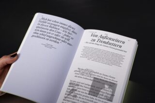 Eine Person hält ein deutschsprachiges Buch aufgeschlagen, auf dessen linker Seite sich ein großes Zitat befindet und auf der rechten Seite eine fettgedruckte Überschrift und der Fließtext zu sehen sind.