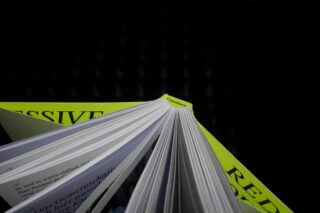 Eine Nahaufnahme eines offenen Buches mit weißen Seiten, das auf zwei geschlossenen Büchern mit hellgrünen Einbänden und teilweise sichtbarem Text vor einem dunklen Hintergrund liegt.