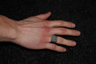 Eine linke Hand mit einem silbernen gemusterten Ring am Mittelfinger ruht auf einer schwarzen strukturierten Oberfläche.