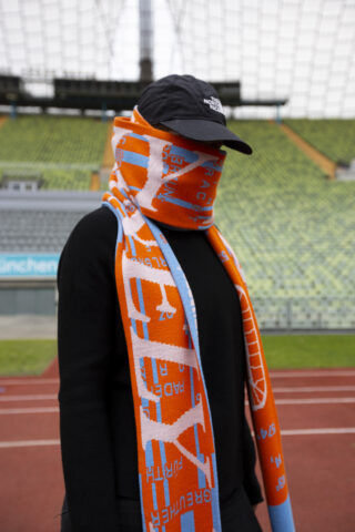 Eine Person mit schwarzer Mütze und Pullover bedeckt ihr Gesicht mit einem orange-blauen Schal und steht auf einer Stadionlaufbahn mit leeren grünen Sitzen im Hintergrund.