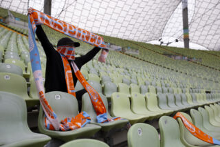 Eine Person in dunkler Kleidung sitzt allein auf grünen Stadionsitzen und hält orangefarbene und blaue Schals hoch und trägt diese. Auf den Sitzen in der Nähe sind weitere Schals drapiert.