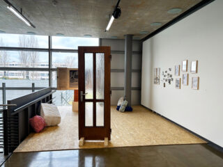 Eine Holztür steht aufrecht auf einem Podest, daneben liegen Kissen; an der angrenzenden weißen Wand hängen gerahmte Fotos und Kunstwerke in einem modernen, offenen Galerieraum.