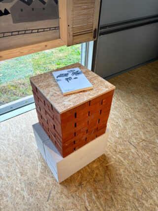 Auf einem Stapel brauner und weißer Ziegelsteine mit einem Holzbrett darauf liegt eine Broschüre in einem Raum mit Holzboden und einem Fenster in der Nähe.