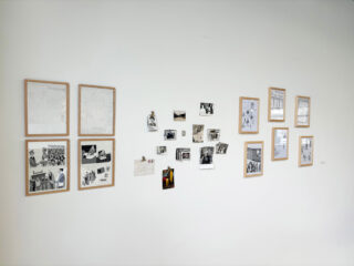 An einer weißen Wand hängen gerahmte Zeichnungen und mehrere kleine Schwarzweißfotos, die in einem lockeren, geordneten Muster angeordnet sind.