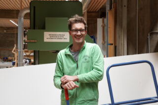 Ein Mann mit Brille und grünem Kapuzenpullover mit Reißverschluss steht lächelnd in einem Werkstattgebäude und stützt sich auf einen Besenstiel.