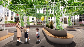 Eine Frau und drei kleine Kinder stehen auf einem modernen Platz im Freien mit geschwungenen Bänken und grünen, belaubten Dachkonstruktionen. In der Nähe schwebt ein gelber Ballon.