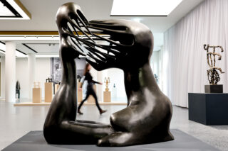 In einer modernen Galerie ist eine große abstrakte Bronzeskulptur einer sitzenden Figur mit langgezogenen, ineinander verschlungenen Gesichtszügen ausgestellt, im Hintergrund geht eine Person vorbei.