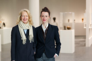 Zwei Frauen in Geschäftskleidung stehen in einer Kunstgalerie, hinter ihnen sind in Glasvitrinen Skulpturen ausgestellt.