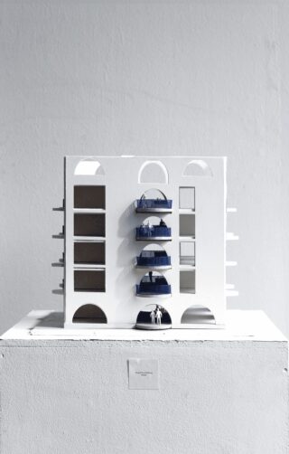 Ein weißes Architekturmodell eines mehrstöckigen Gebäudes mit Balkonen und kleinen blauen menschlichen Figuren wird auf einem weißen Sockel vor einer schlichten grauen Wand angezeigt.
