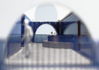 Ein Miniatur-Architekturmodell zeigt abstrakte menschliche Figuren in einem blau-weißen gewölbten Innenhof mit gefliesten Wänden und offenen Durchgängen.